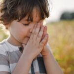 Little Boy closed her eyes, praying in a field wheat. Hands folded in prayer. Religion concept